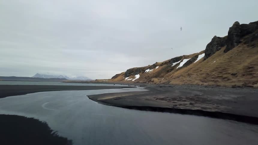Drone footage shot just above the surface following a river leading to a larger body of water on a cloudy day