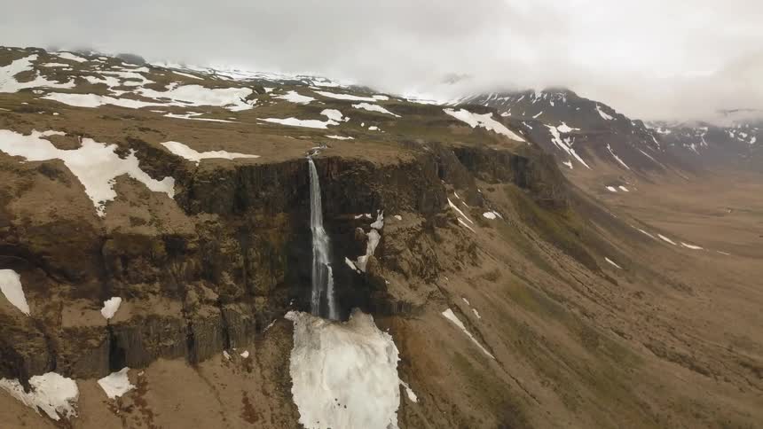 A waterfall is flowing over a cliff onto a patch of ice on a mountainside during the day