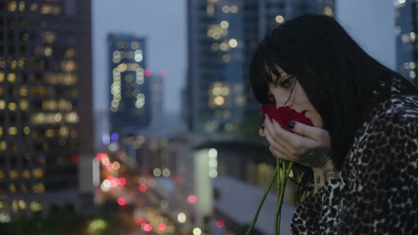 A woman stands atop a building cupping a rose to her face