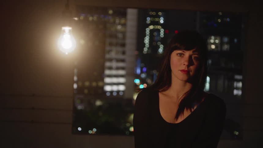 Woman on a balcony in a large city at night next to a lighted bulb