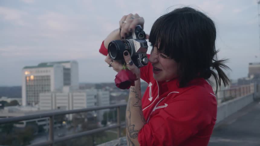 A woman is holding a rose and taking pictures as she stands on a rooftop at dusk