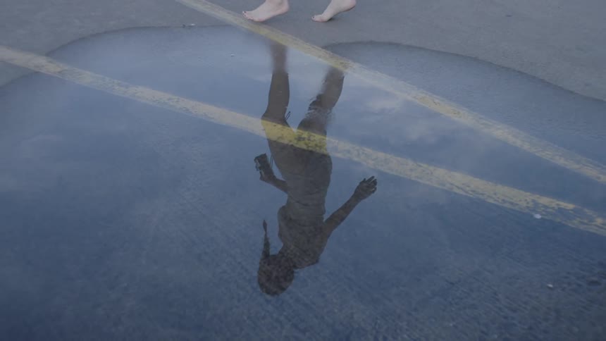 A barefooted woman is walking around a puddle on an asphalt surface in daylight