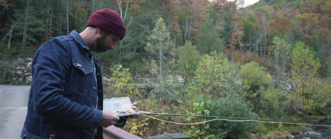 A man is looking in a box containing lures as he fishes from a bridge during the day