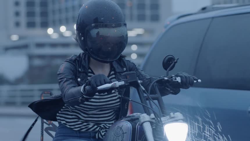 A woman is riding a motorcycle on a city street at dusk in the rain