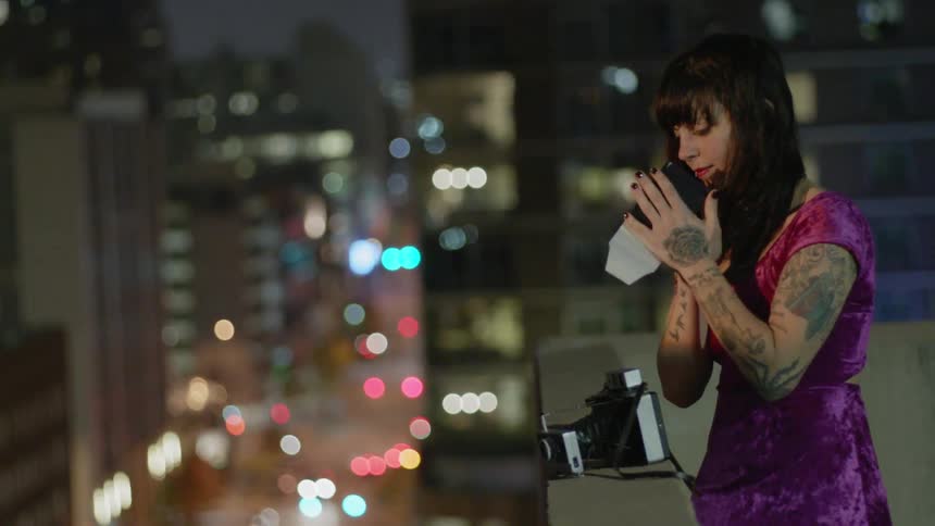 A woman is shaking a polaroid picture as she stands on a rooftop at night