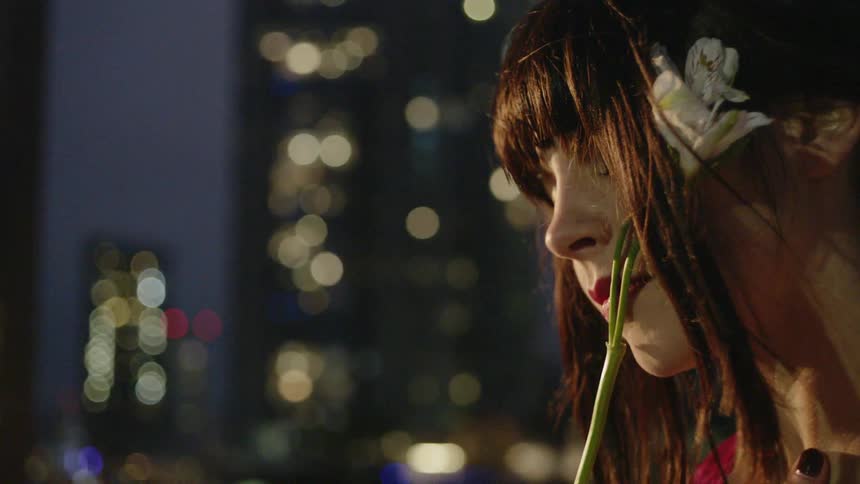A woman holds a flower next to her face on a city rooftop