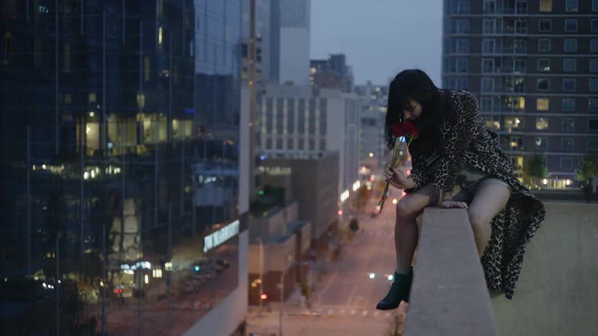 A woman hangs over the ledge of a roof holding a red rose