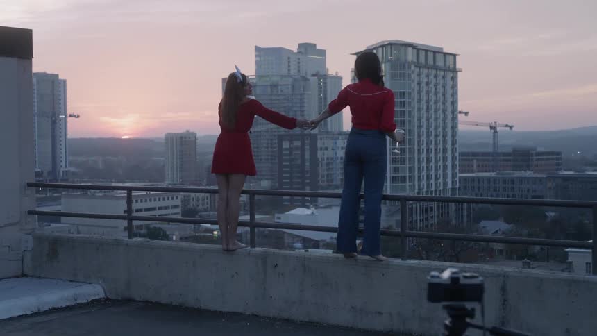 Two women are holding hands as they stand on the edge of a rooftop at dusk