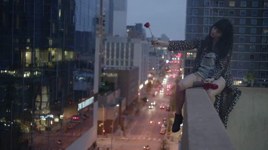 A girl is sitting on the ledge of a large building dangling a flower over the edge