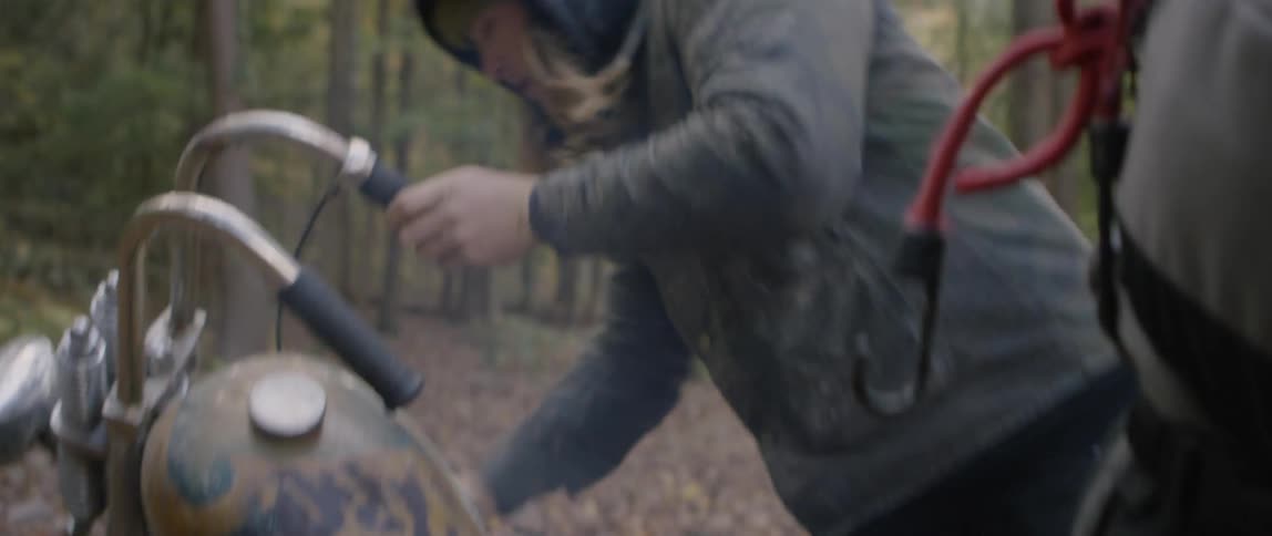 A man is trying to kickstart his motorcycle outside in a forest during the day