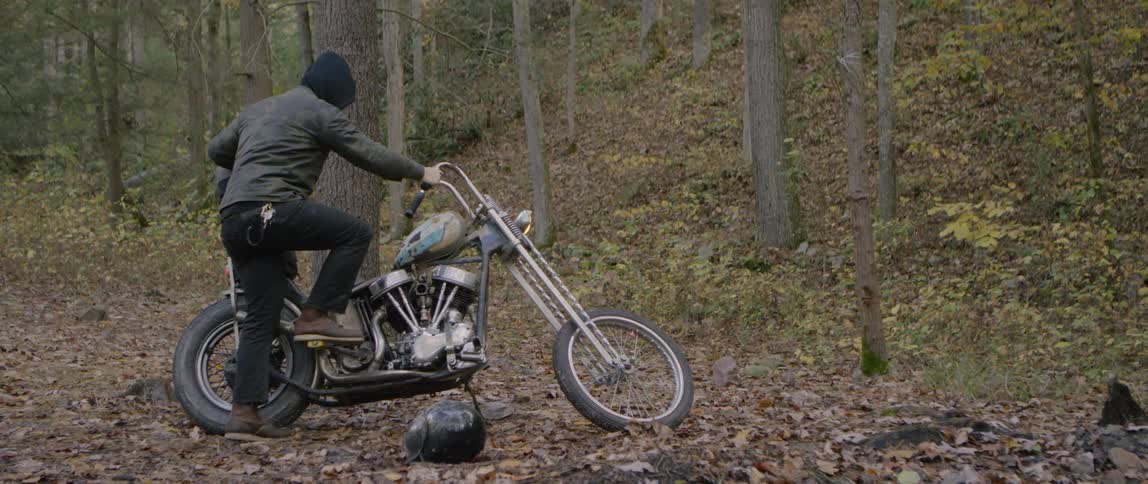 A man is trying to kick start his motorcycle near the woods