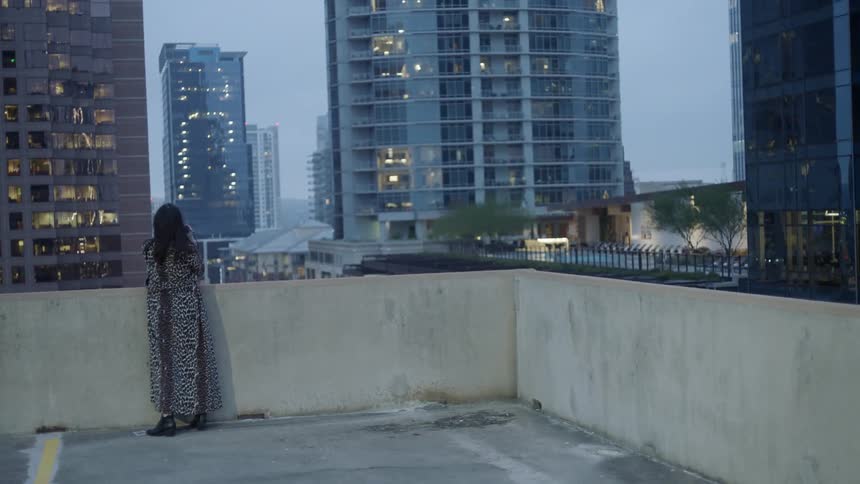 A woman stands on a rooftop looking out at the city buildings