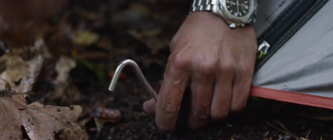 A man is using a rock to pound a tent stake into the ground