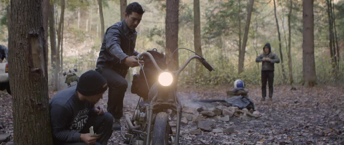 A man is trying to kickstart a motorcycle outside in a forest campsite during the day