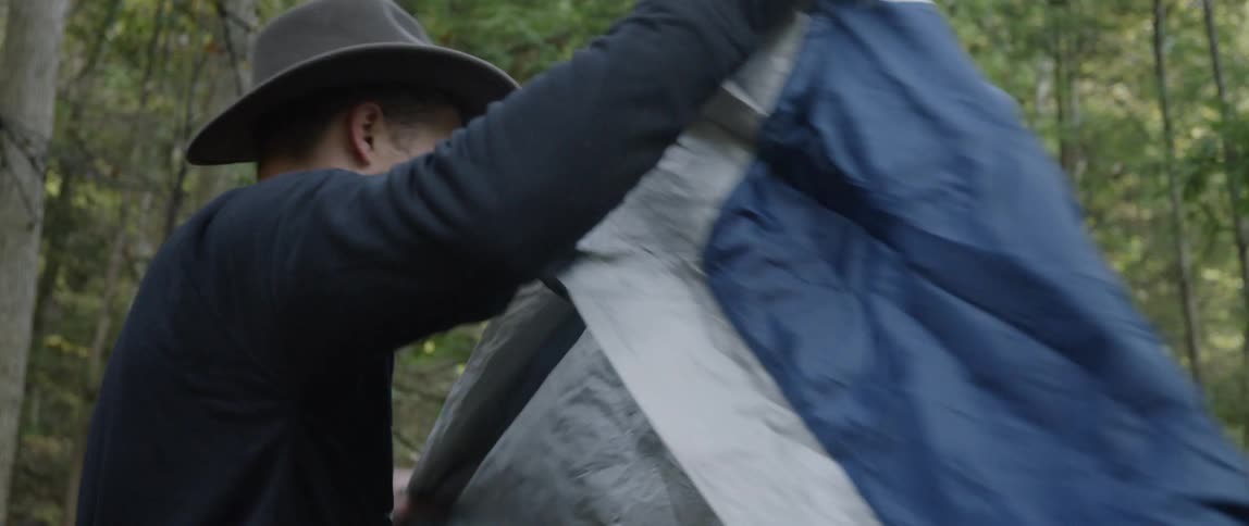 A man is unfolding a tent as he stands in a forest during the day