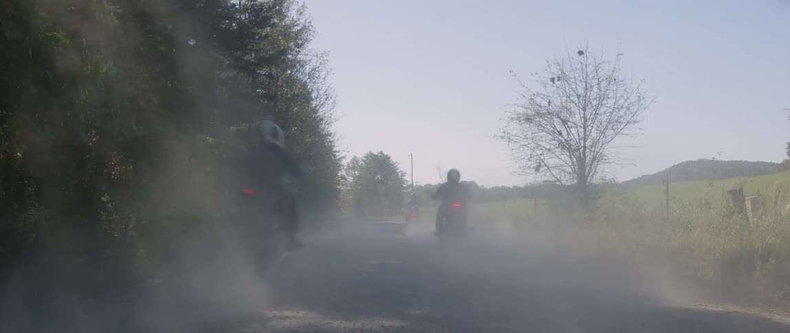The motorcycles race past the field and forest on a sunny day