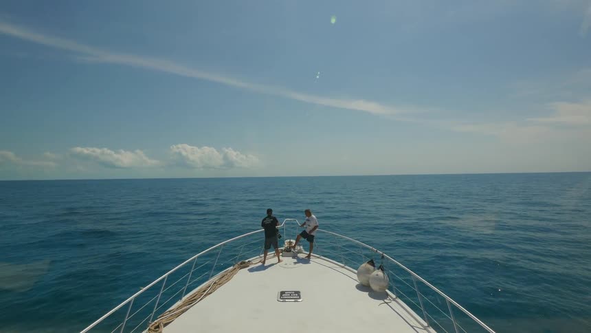 Two men are standing on the bow of a boat as it sails on a sunny day