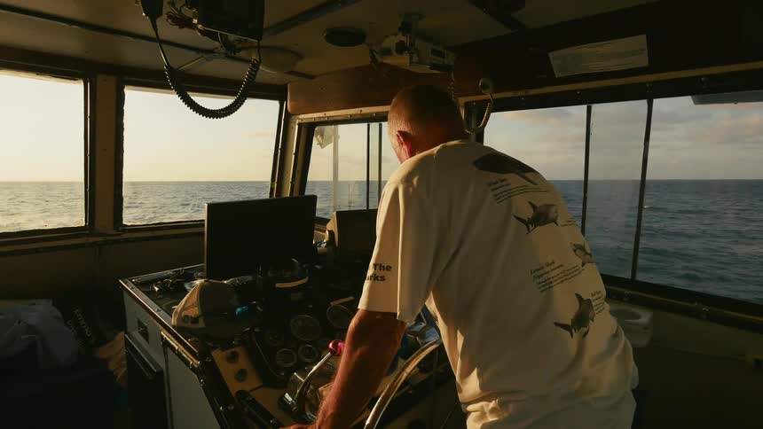 A man gets up to stand at the wheel of a boat as it sails in the afternoon