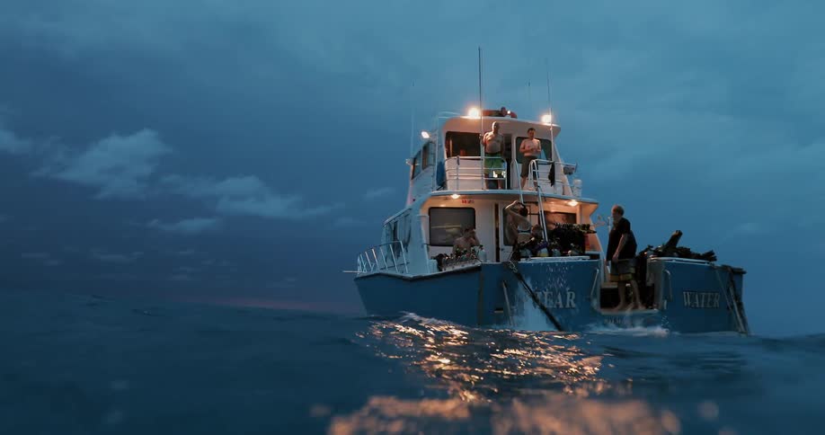 A double decker boat with several people on it sails on choppy water at dusk