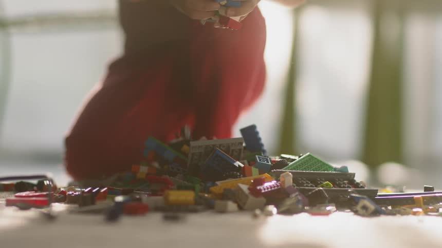 A boy is picking up and dropping bunches of building blocks into a pile in a room during the day