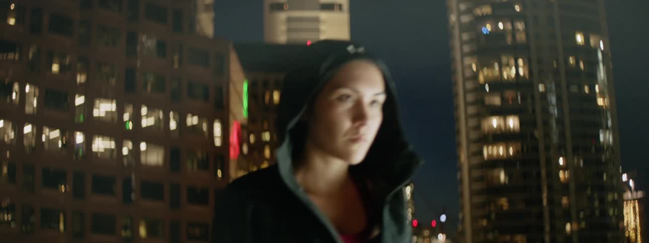 A woman puts her hood up and walks away in a city at night