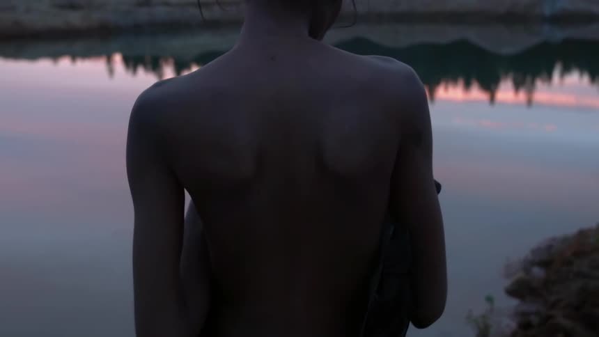 A shirtless person stands next to a lake at sunset