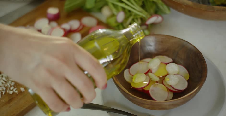 A woman drizzles olive oil over radishes in a wood bowl