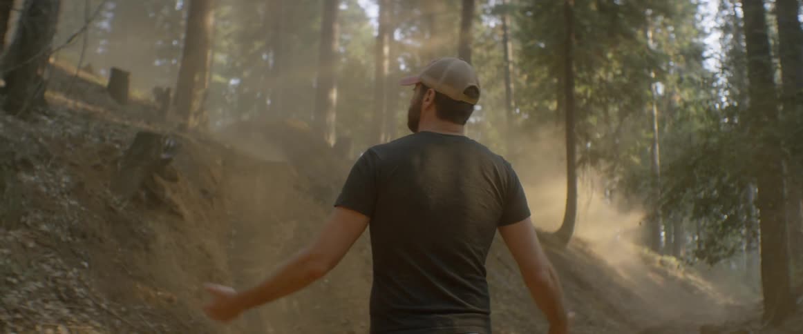 A man is shrugging as he looks at dust trailing away in a forest during the day