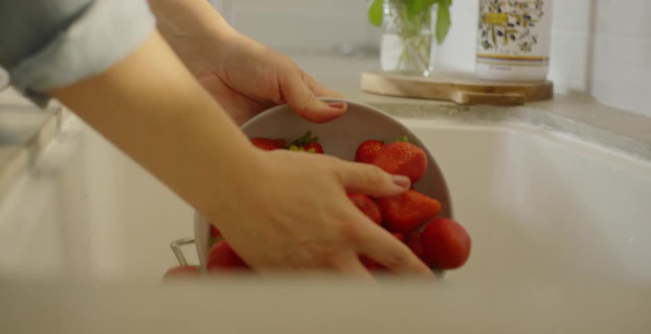 A woman puts a bowl of red strawberries into a bowl in the sink