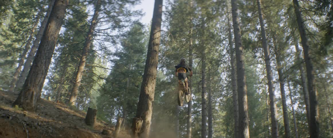 A man is watching a woman jump a motorcycle on a hill in a natural setting