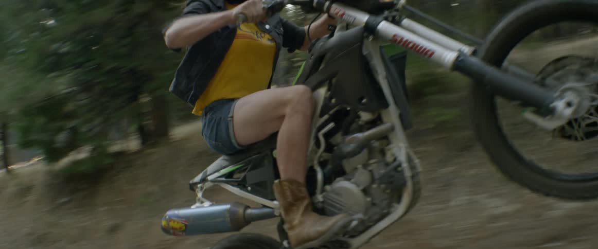 A woman pops a wheelie on a motorcycle in a forest