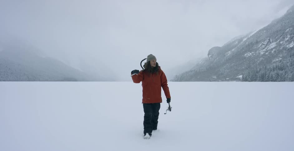 A man is walking across a frozen lake carrying a chainsaw and fishing pole