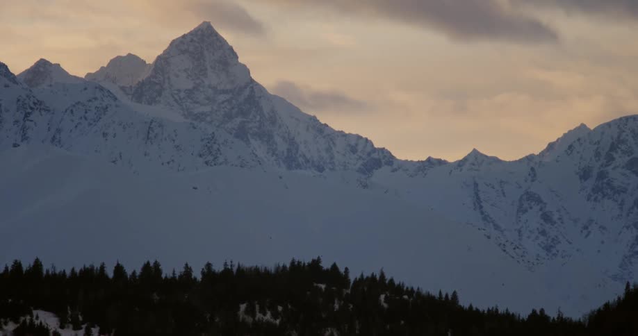 Clouds are moving across snow-covered mountains at dusk