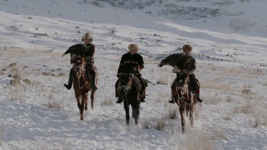 Three people ride horses over the snow and two of them carry birds on their arms