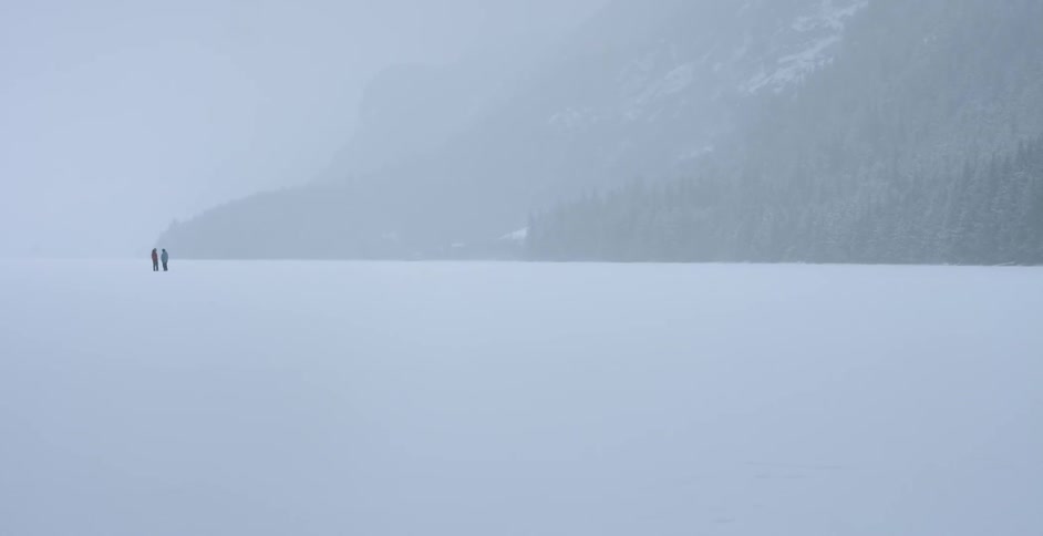 Two people standing in a large open snow-covered area next to a forest and some mountains