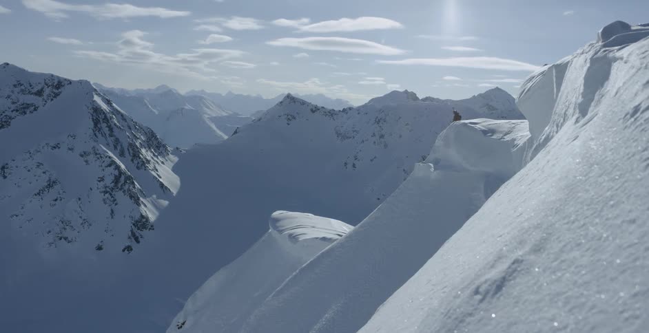 A person is sitting on a snowy cornice overlooking a mountain range during the day