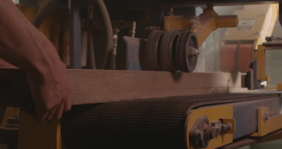 A white man feeds a board through a machine and another worker pulls it through