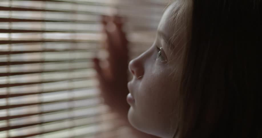 A young girl looks out of window blinds