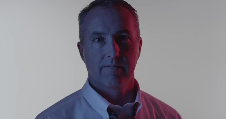 A man with purple and pink light on his face is looking at the camera