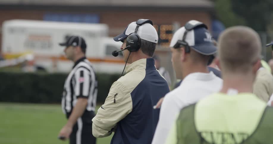 A football coach is watching the game from the sideline with a headset