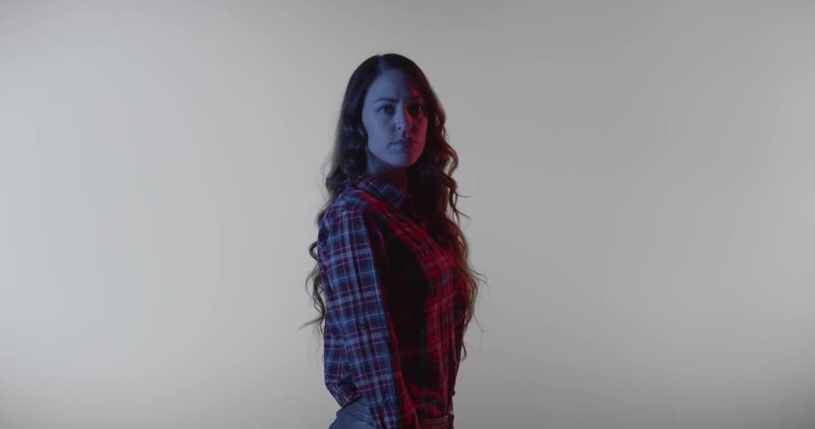 A woman poses and turns in a dim room with a white backdrop