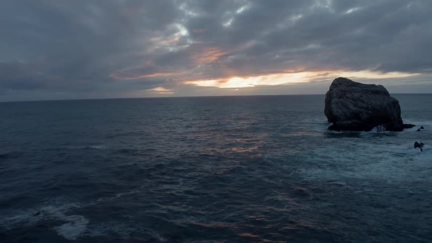 A large rock is sticking out of the sea as the sun sets