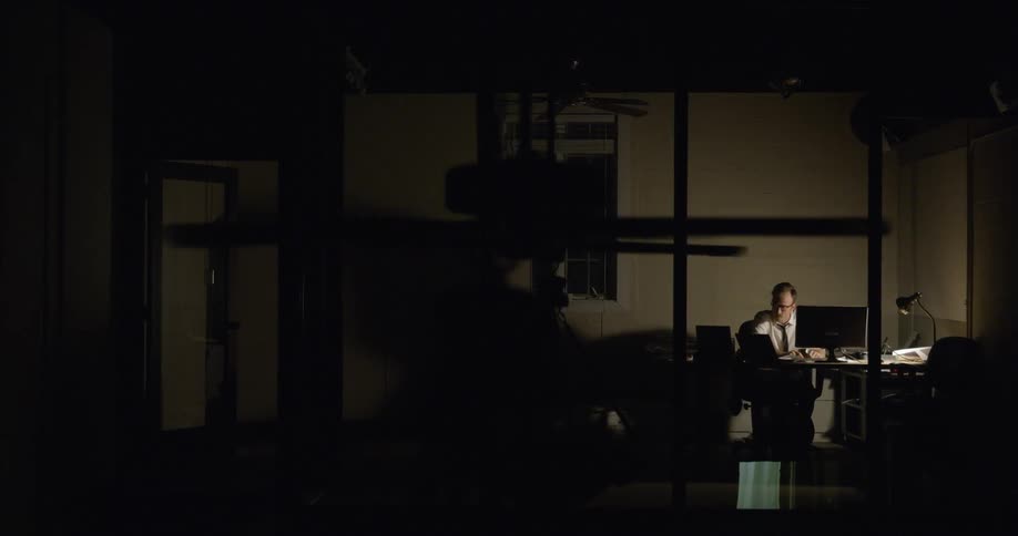 A man sits at a desk in a dark office room as a ceiling fan spins