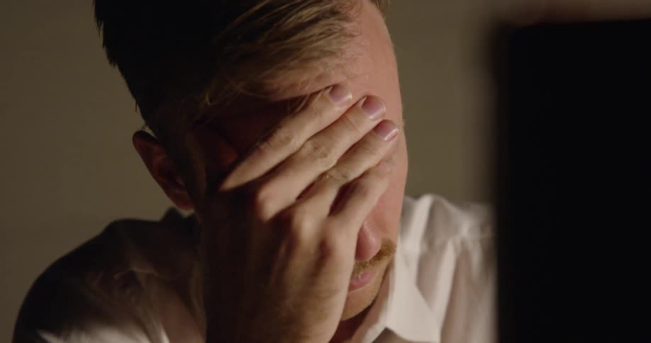 A man sits at a desk and rubs his eyes
