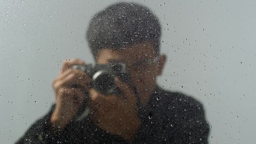 Bespectacled man behind a piece of glass with droplets of water on it raises his camera and takes a picture