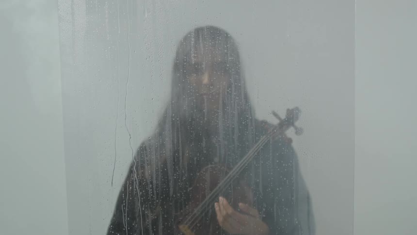 Person standing and holding a small stringed instrument behind a pane with water dripping down it