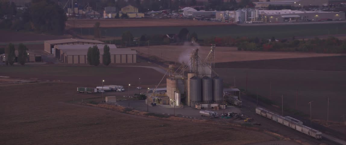 Smoke is pouring from a stack at a silo complex in an agricultural setting at dusk
