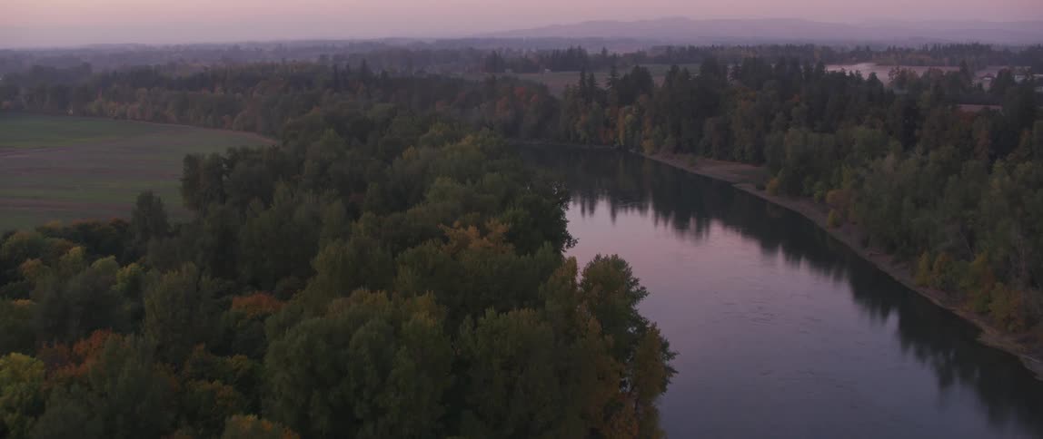 A river is flowing past fields and forest trees at dusk