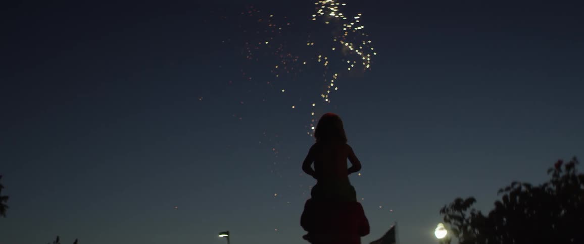 A girl sits on her parent's shoulders as fireworks go off