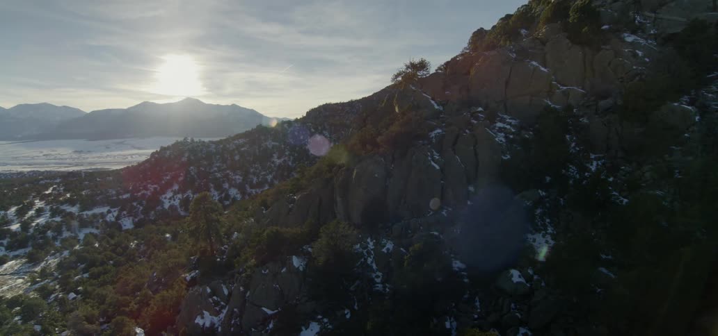 The sun shines on the side of a partially snow covered mountain